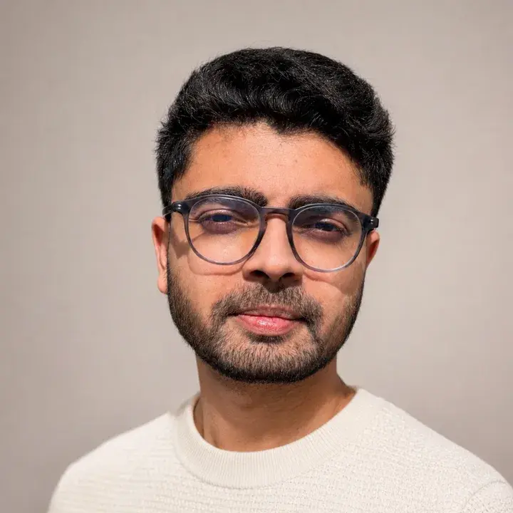 Portrait of Ravi, with short dark hair, a beard and round glasses, wearing a cream knit sweater against a plain wall.