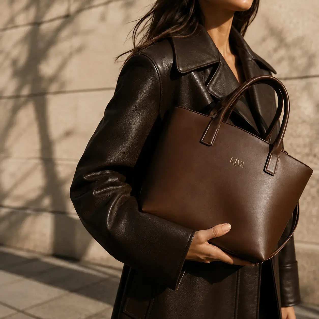 Model holding a brown RIVA handbag while wearing a dark brown coat in warm sunlight.