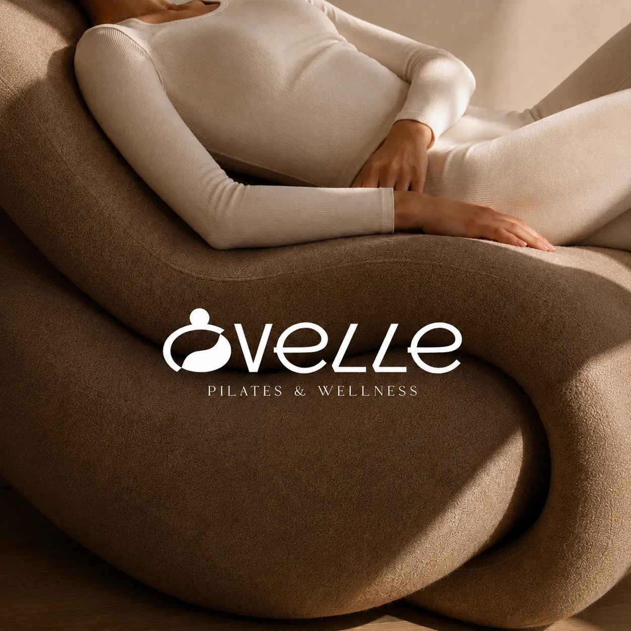 Ovelle Pilates and Wellness wordmark over a warm brown sculptural lounge chair and neutral activewear.