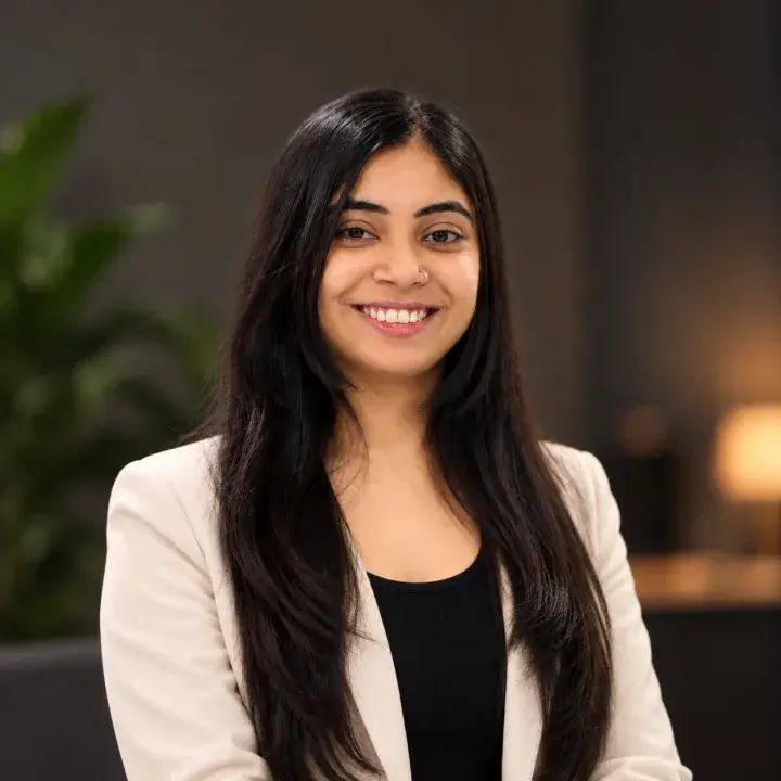Portrait of Aarti, smiling, with long dark hair, wearing a cream blazer over a black top, in a warmly lit room with a plant and a lamp behind her.